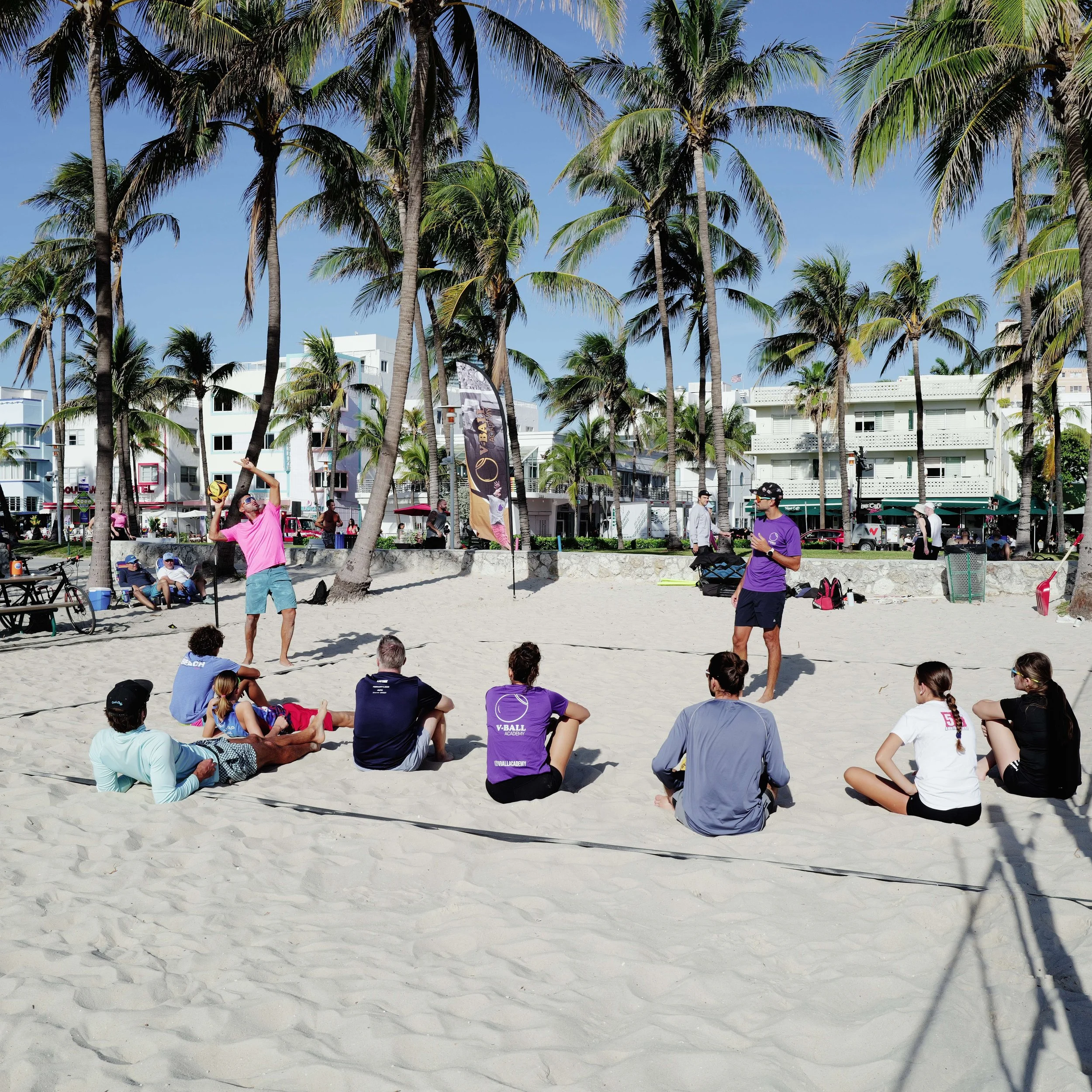 People sitting on the sand at a beach volleyball lesson, with a coach demonstrating a volleyball technique while others watch, surrounded by palm trees and beachside buildings in the background.