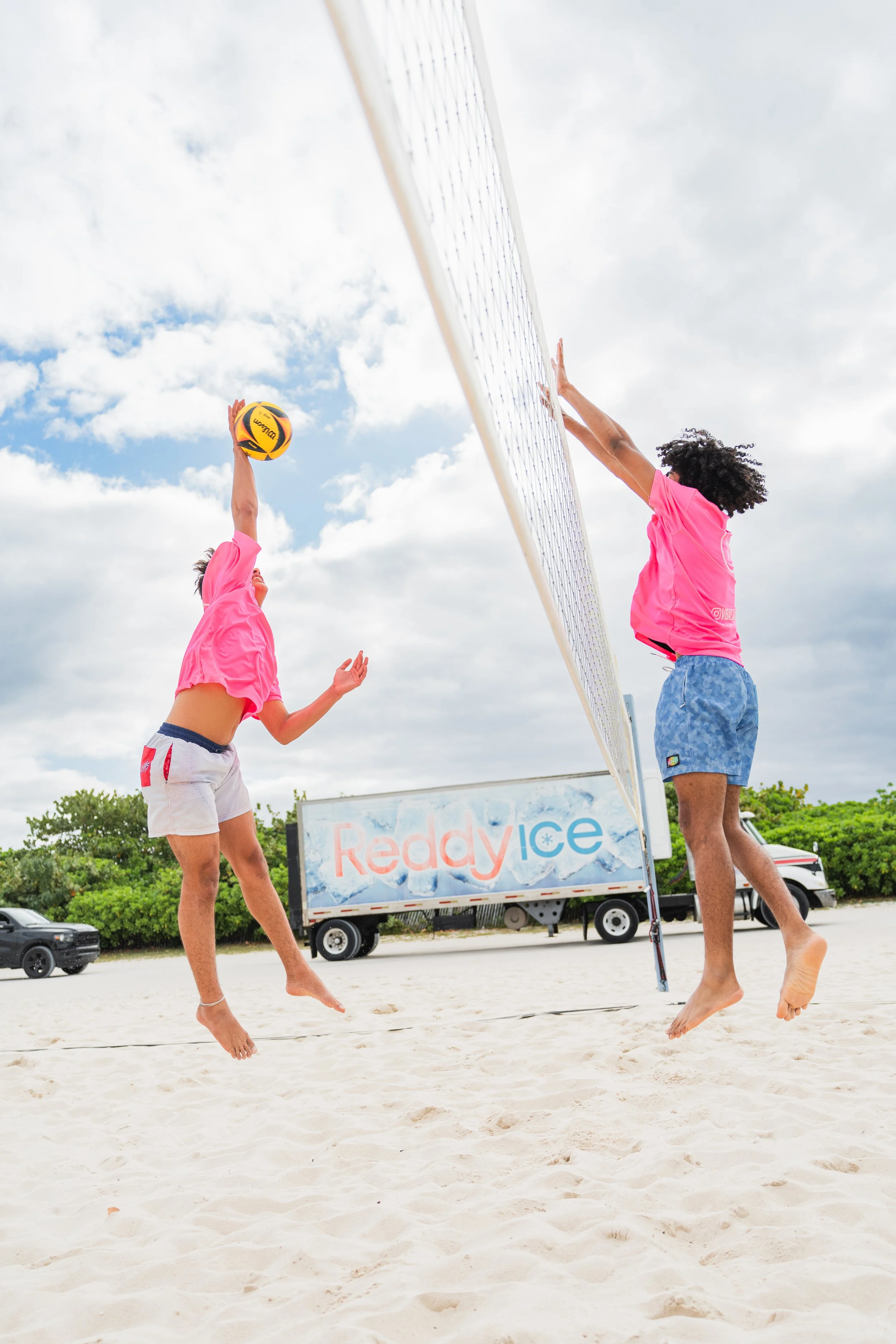 Two women playing beach volleyball on sand, one jumping to hit the ball, the other jumping to block it, with a sign that says 'Reddy Ice' in the background, and vehicles parked behind trees under a cloudy sky.