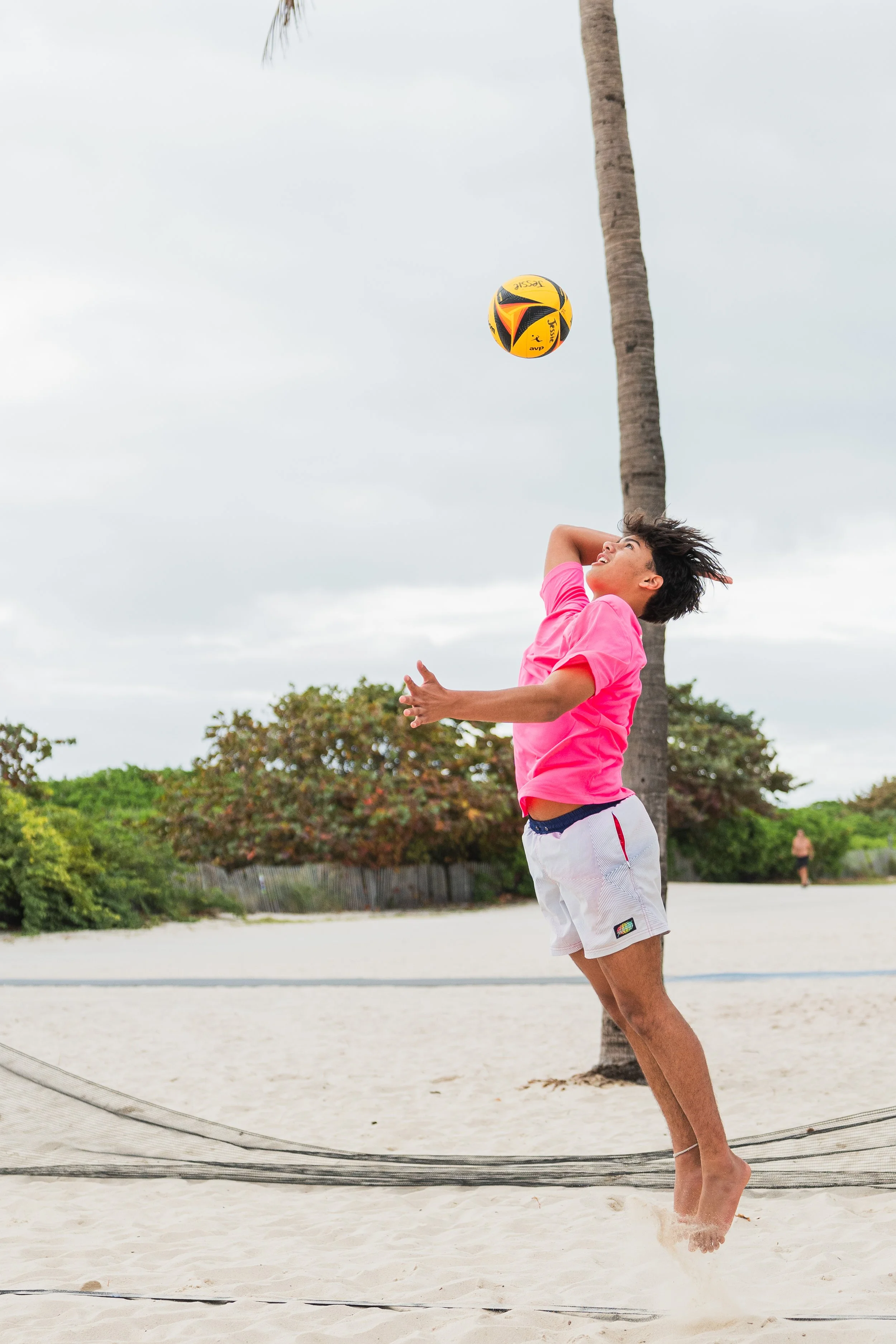 A young person in a bright pink shirt and white shorts is playing volleyball on a sandy beach. They are jumping with their arm raised, about to hit the yellow and black volleyball. A palm tree is in the background, along with some green bushes and an overcast sky.