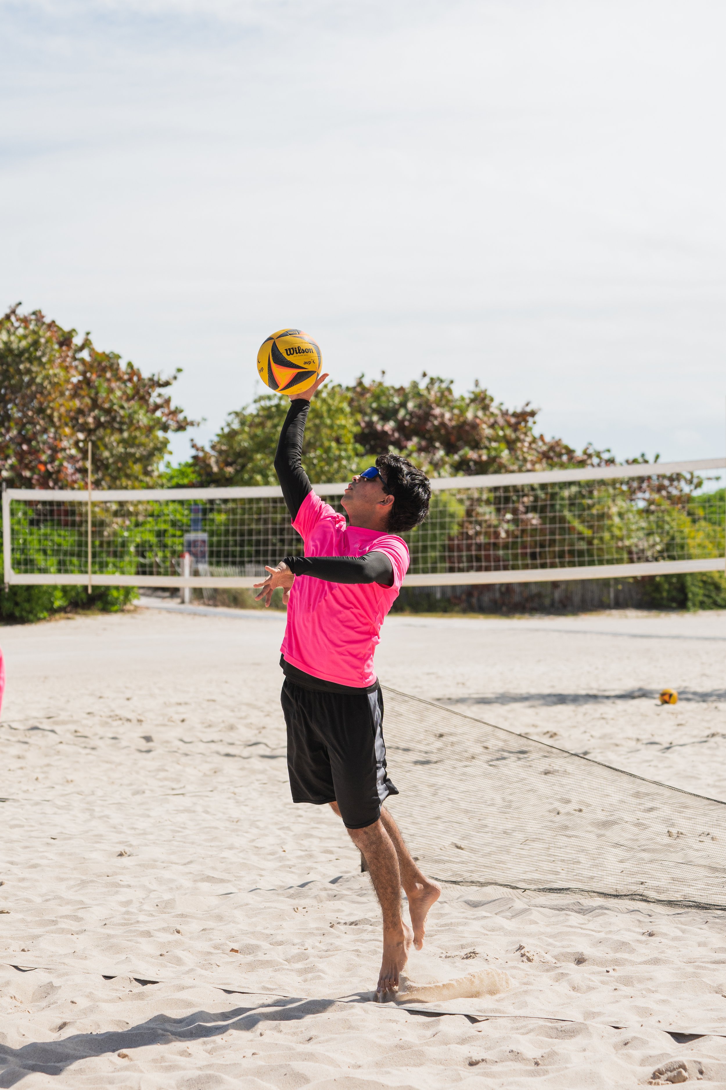 A man playing beach volleyball, jumping to hit a yellow volleyball on a sandy court with a net and trees in the background on a partly cloudy day.