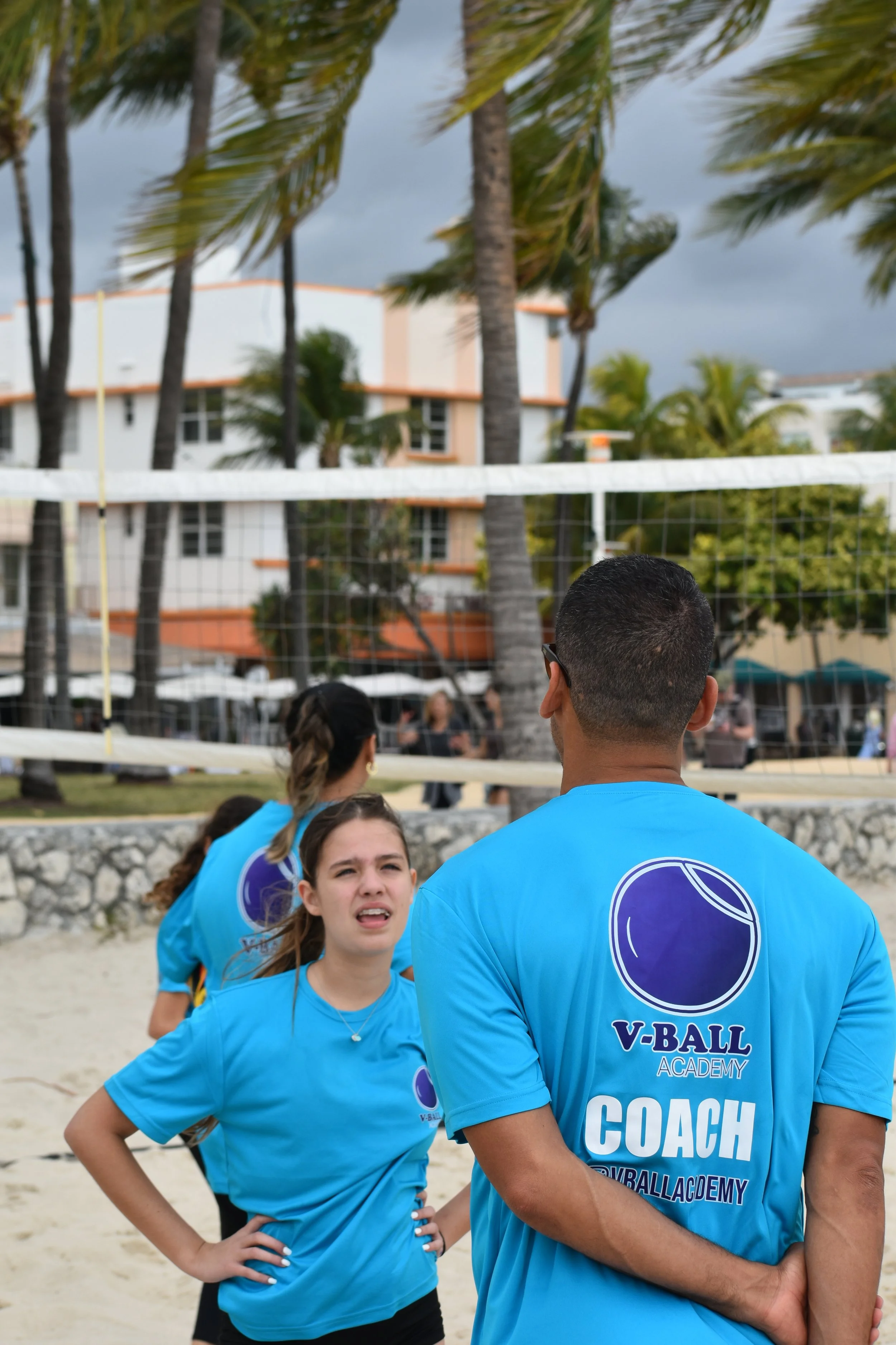 Beach volleyball coach and players wearing blue shirts with V-BALL Academy logo, standing on sandy court with volleyball net, palm trees, and buildings in the background.