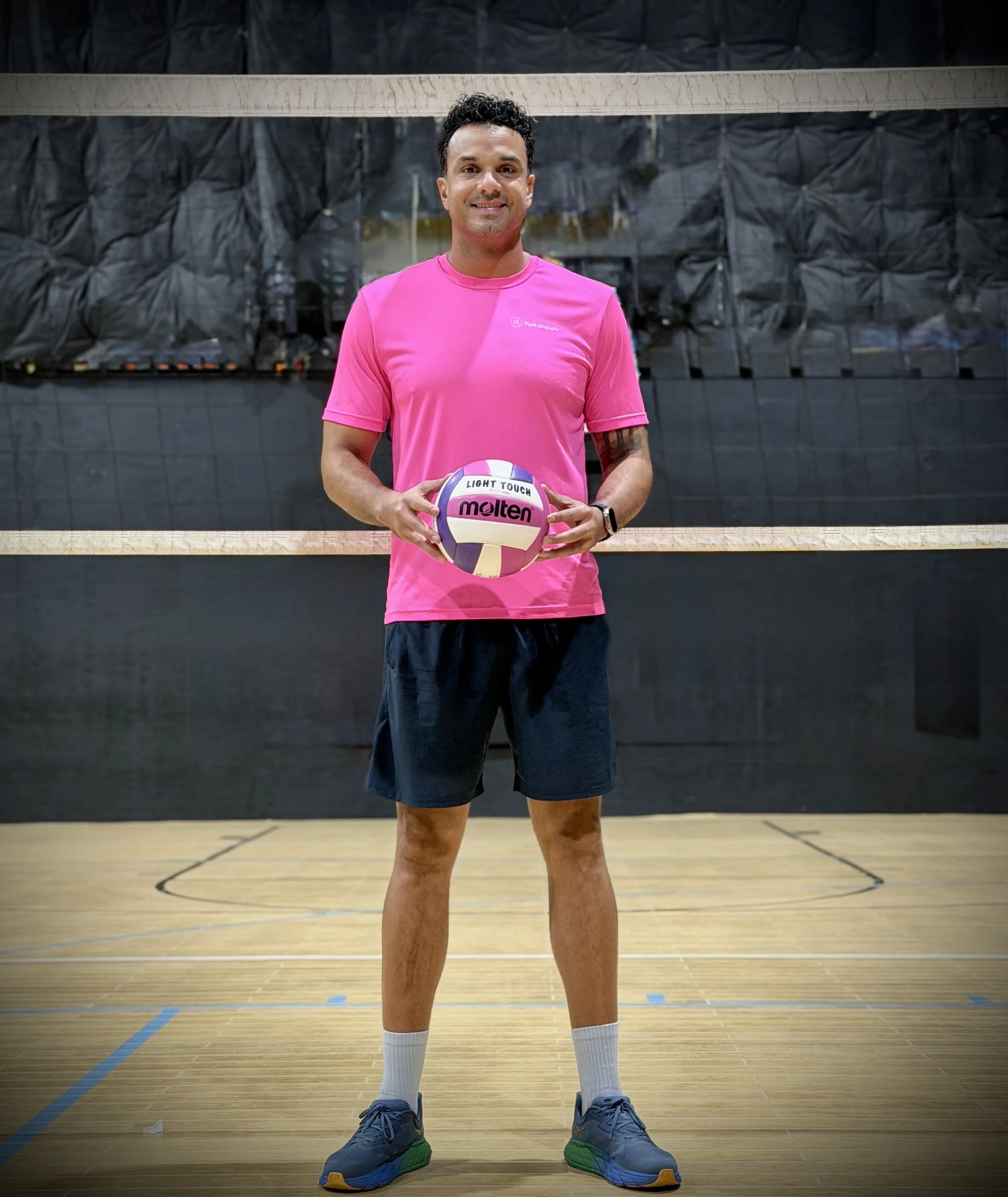 A man in a pink sports shirt and black shorts standing on an indoor volleyball court, holding a volleyball, with a volleyball net in the background.