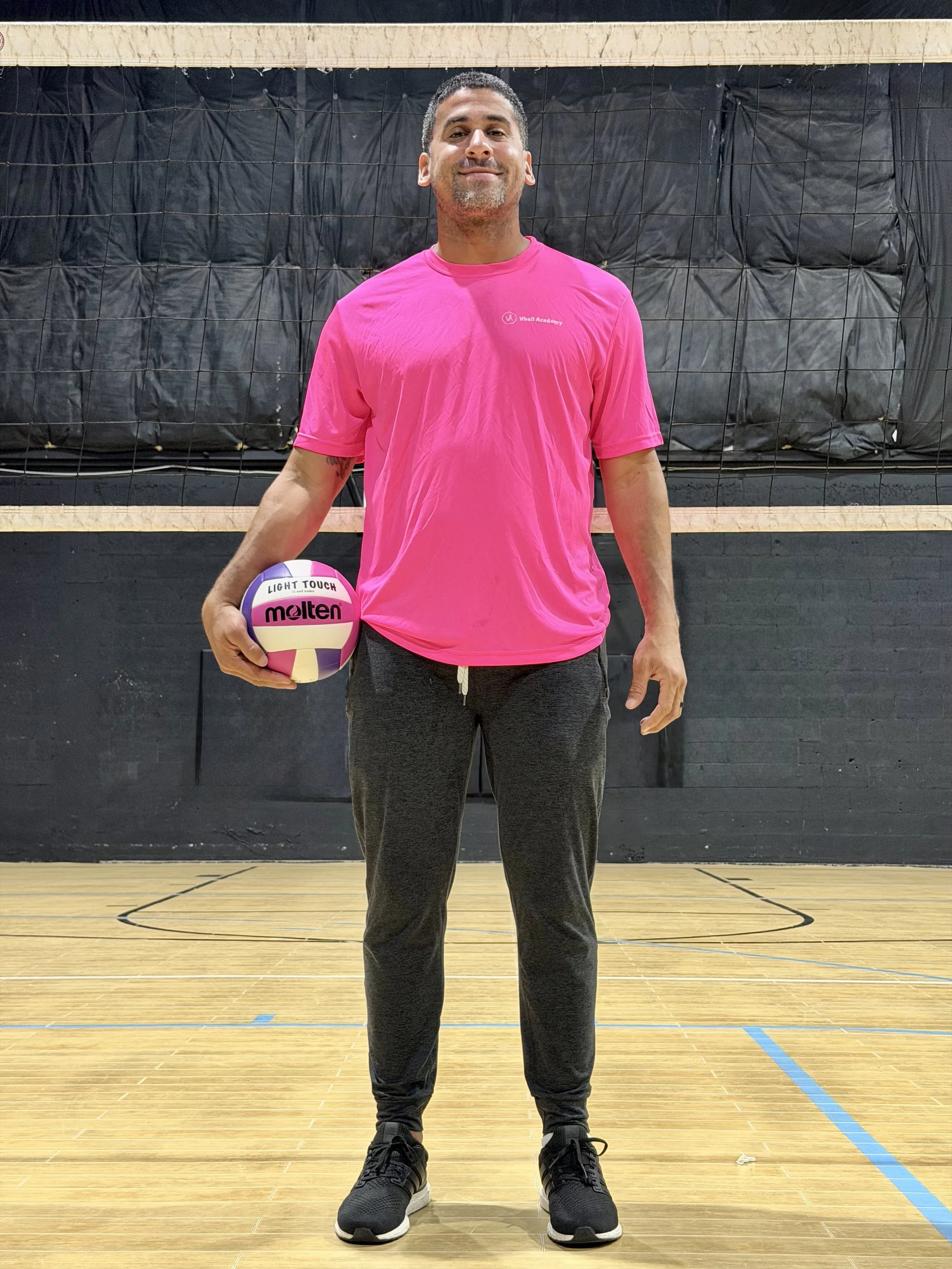 A man in a pink T-shirt and dark gray sweatpants standing on a volleyball court, holding a pink and white volleyball in his right hand, smiling at the camera.