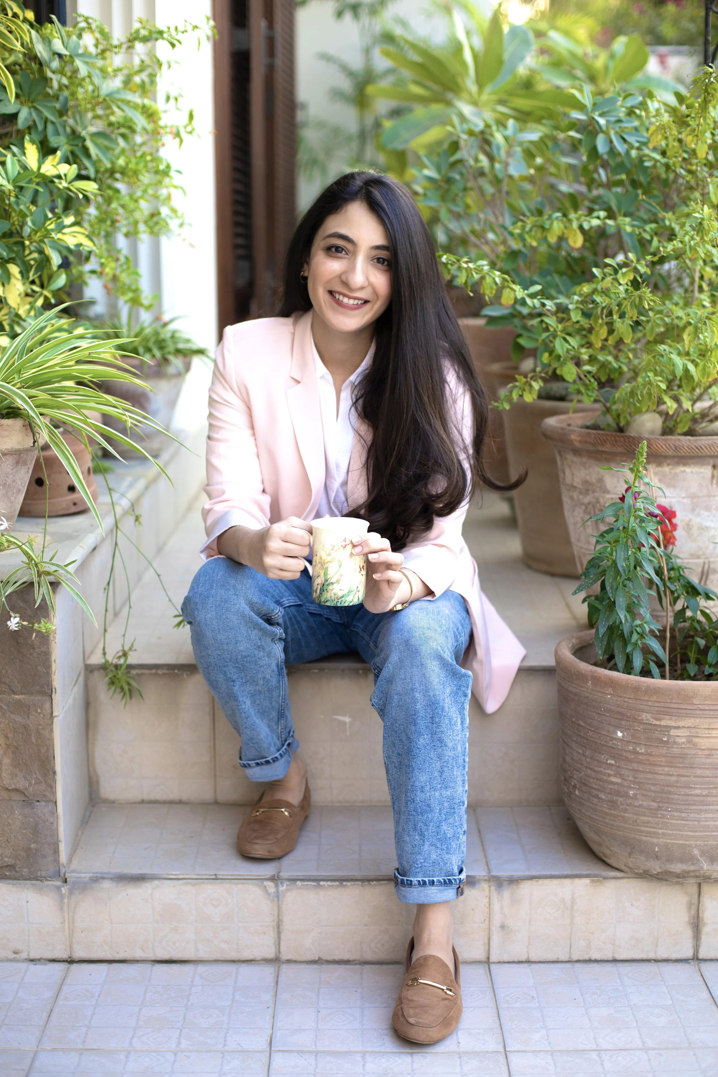 A woman sitting on outdoor steps, holding a cup, surrounded by potted plants, smiling at the camera.