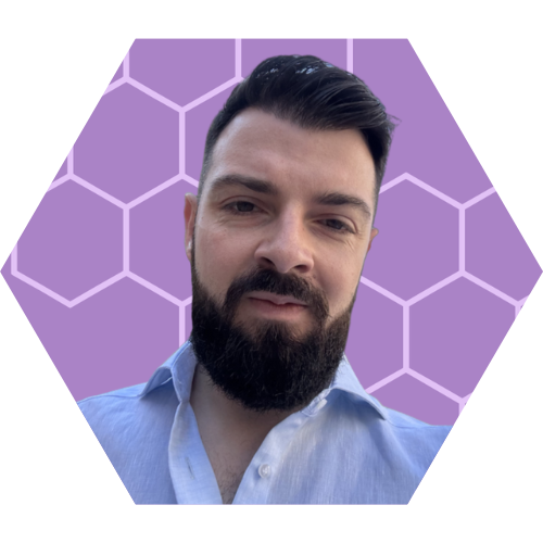 Portrait of a man with dark hair and a beard, wearing a light blue collared shirt, against a purple background with a hexagonal pattern.