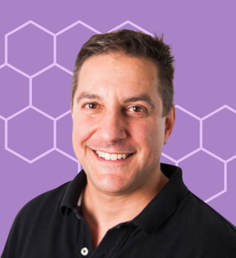 A smiling man with short brown hair wearing a black collared shirt against a purple background with white hexagonal pattern.