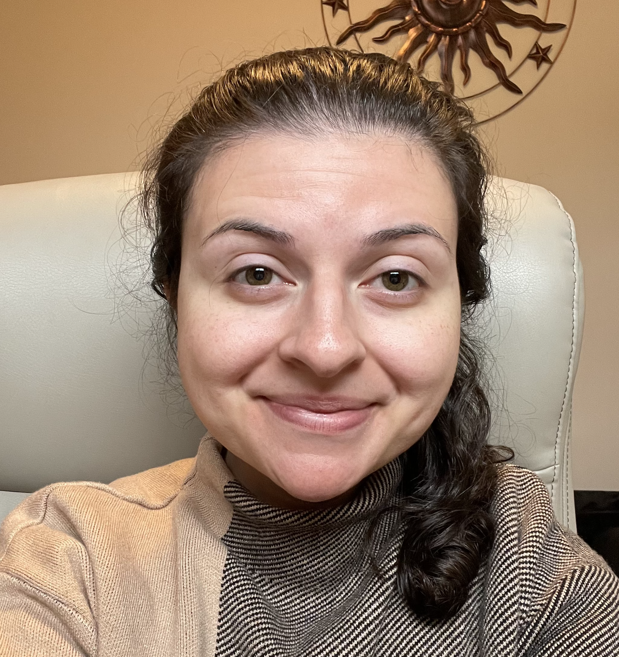 A woman sitting in a chair, smiling at the camera. She has dark curly hair, light skin, and is wearing a brown turtleneck sweater. A decorative sun and star wall art is visible in the background.