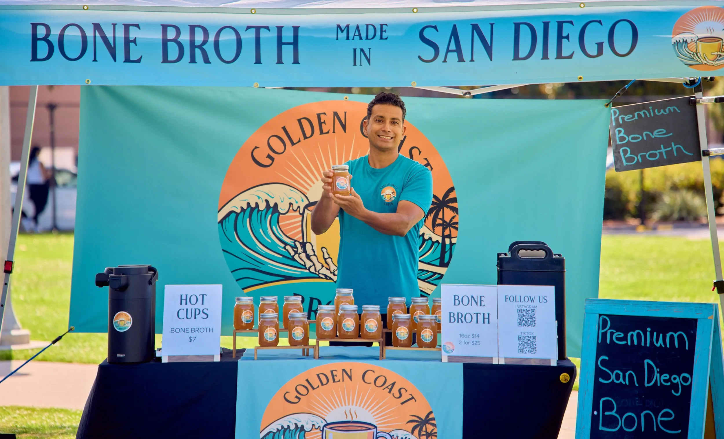 A man standing behind a booth selling bone broth, holding a jar and smiling, with a sign that reads 'GOLDEN COAST BONE BROTH' and prices listed for bottles. The booth has colorful banners and signs advertising the product.