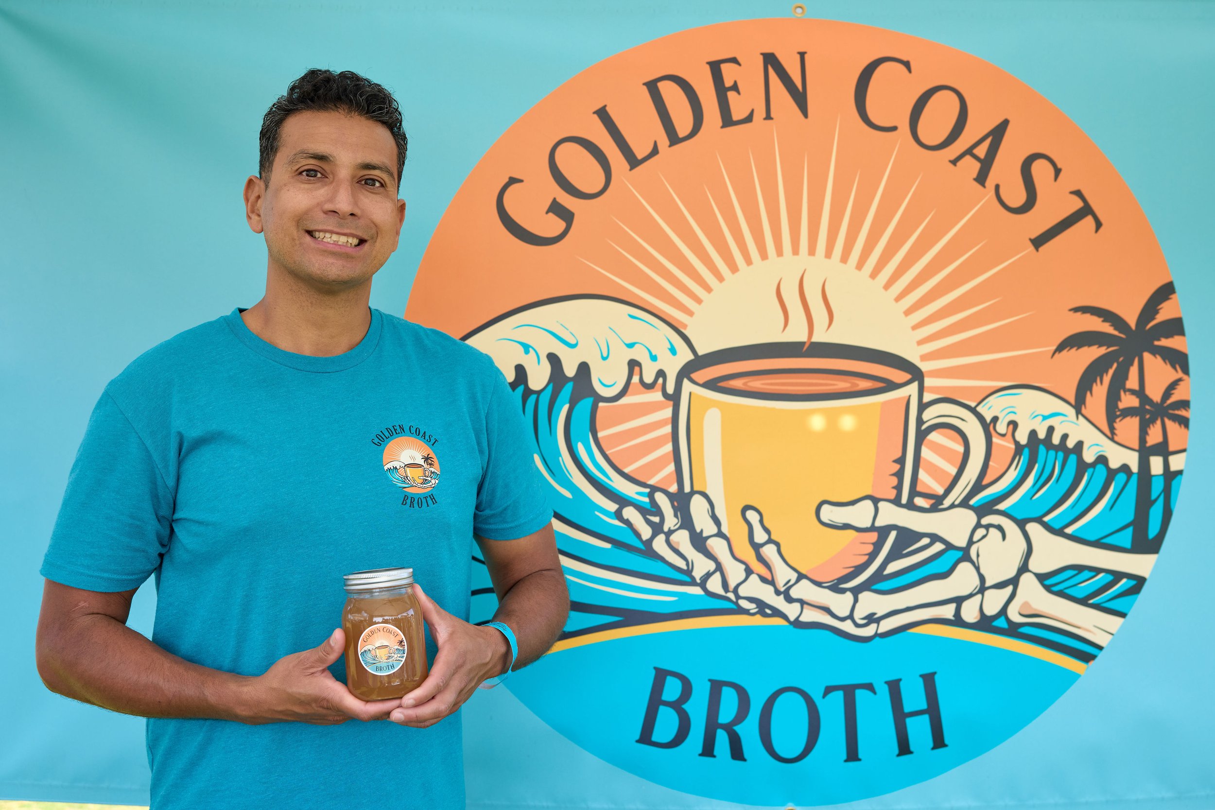 Man wearing a blue t-shirt holding a jar of broth in front of a colorful sign for Golden Coast Broth, featuring a sunrise, waves, a steaming cup, and a palm tree.