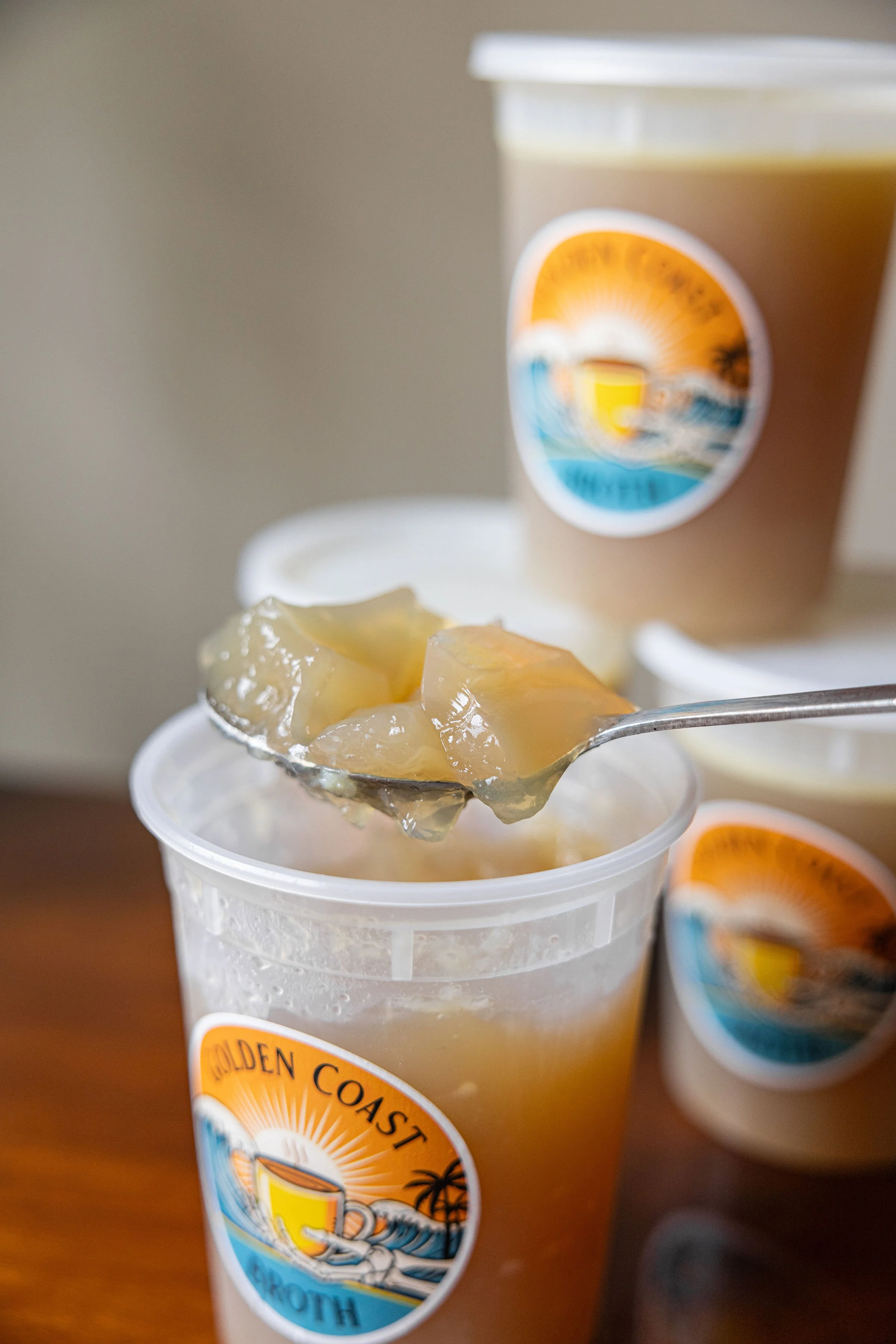 A clear plastic cup with a "Golden Coast" logo, containing gelatinous broth and a spoon. A second similar cup and a lid are in the background.