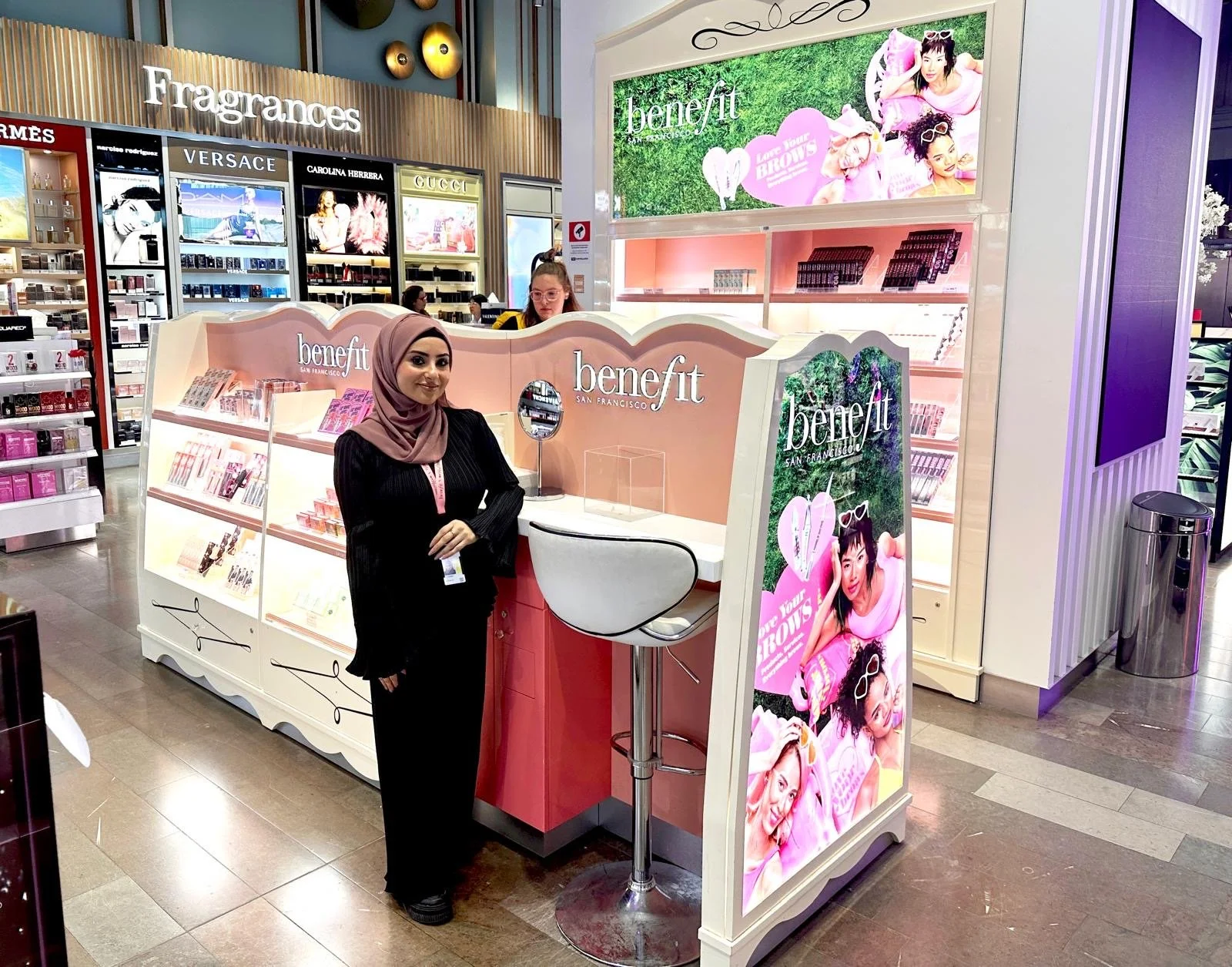 A woman wearing a pink hijab and black outfit standing at a Benefit cosmetics booth inside a store, with various makeup products and advertisements visible in the background.