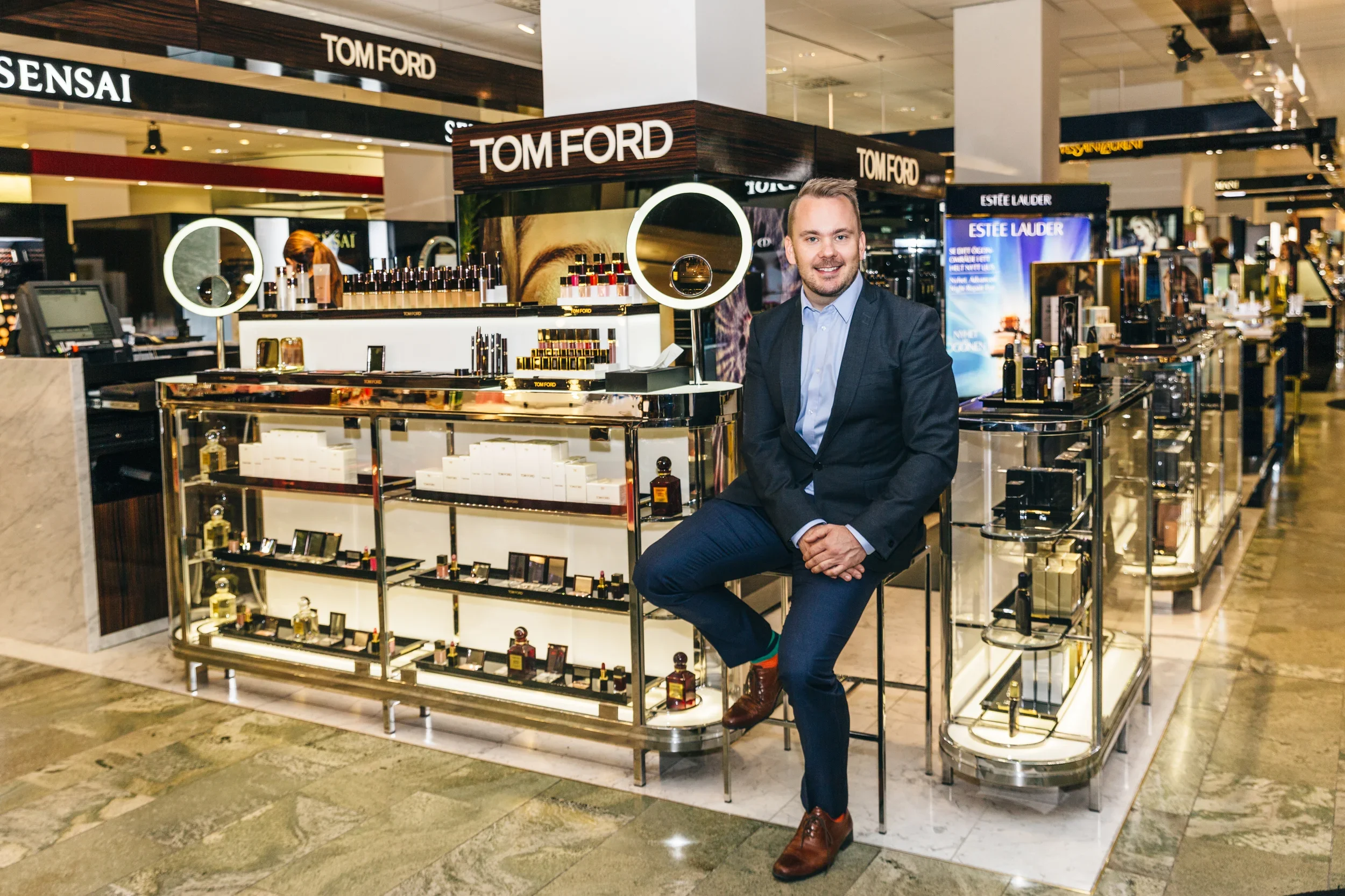 A man sitting on a high stool in front of a perfume display at a shopping mall. The display features Tom Ford perfumes, with bottles and packaging arranged on glass shelves. In the background, there are brands like Sensai and Estée Lauder, and the mall interior is visible.