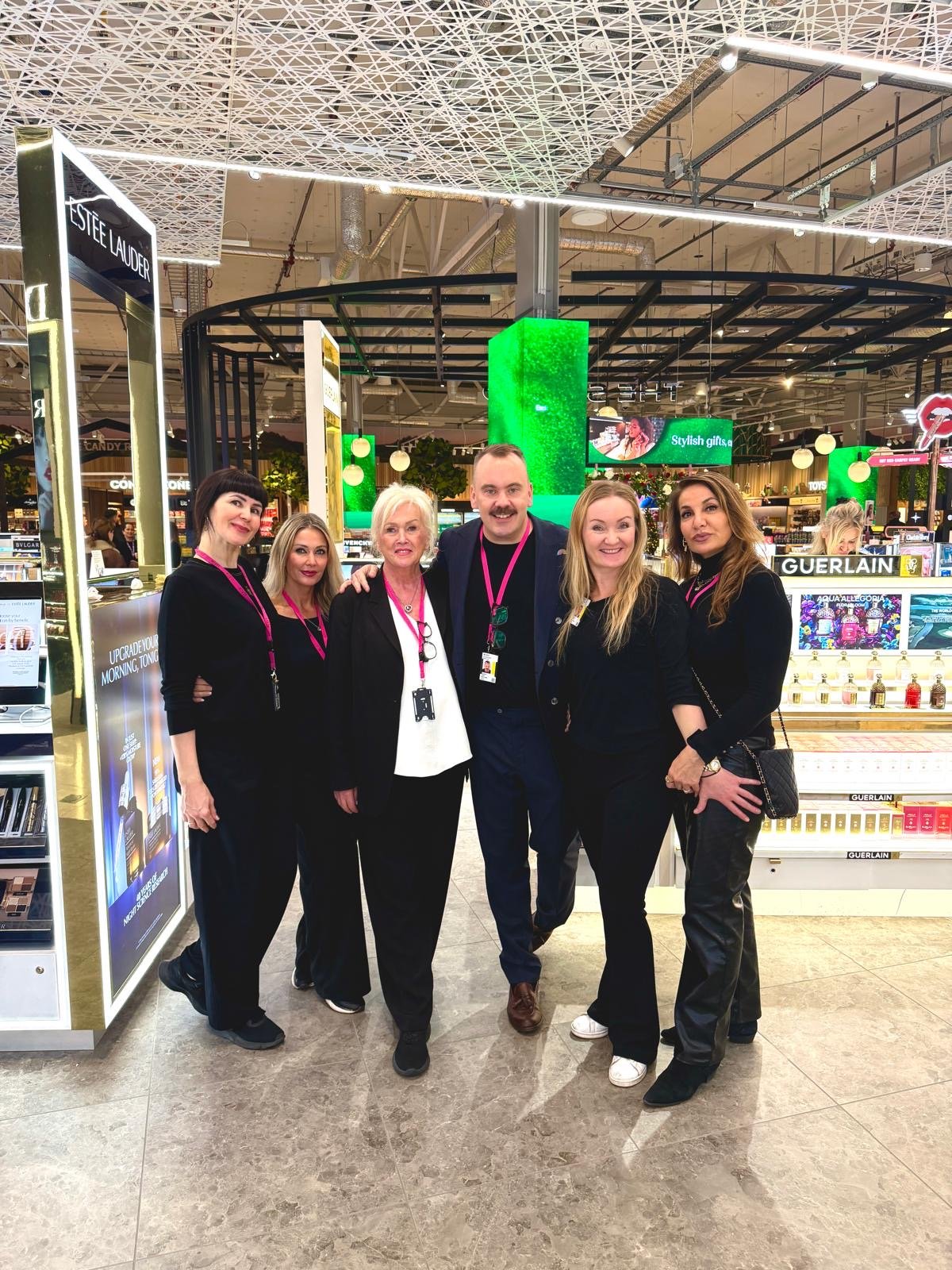 Group of six people standing together in a cosmetics store, smiling at the camera, with shelves of beauty products in the background.