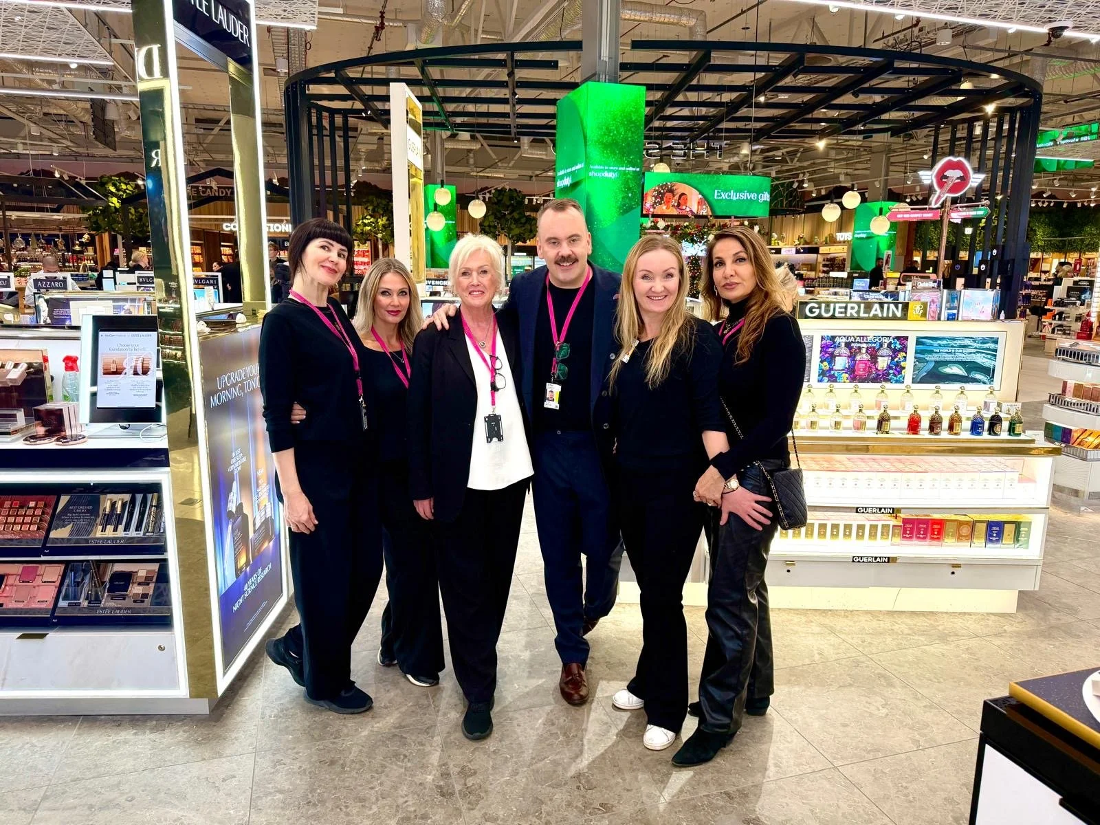 Group of six people posing together inside a store, surrounded by cosmetics and skincare products, with green digital signage in the background.