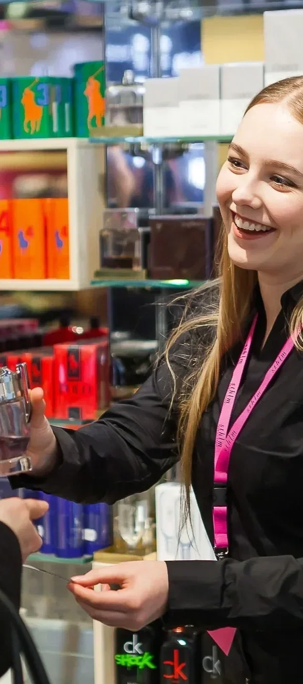 A smiling female cashier with long blonde hair, wearing a black shirt and pink measuring tape around her neck, is friendly handing a receipt or change to a customer at a store counter. Shelves behind her are stocked with colorful boxes and bottles.