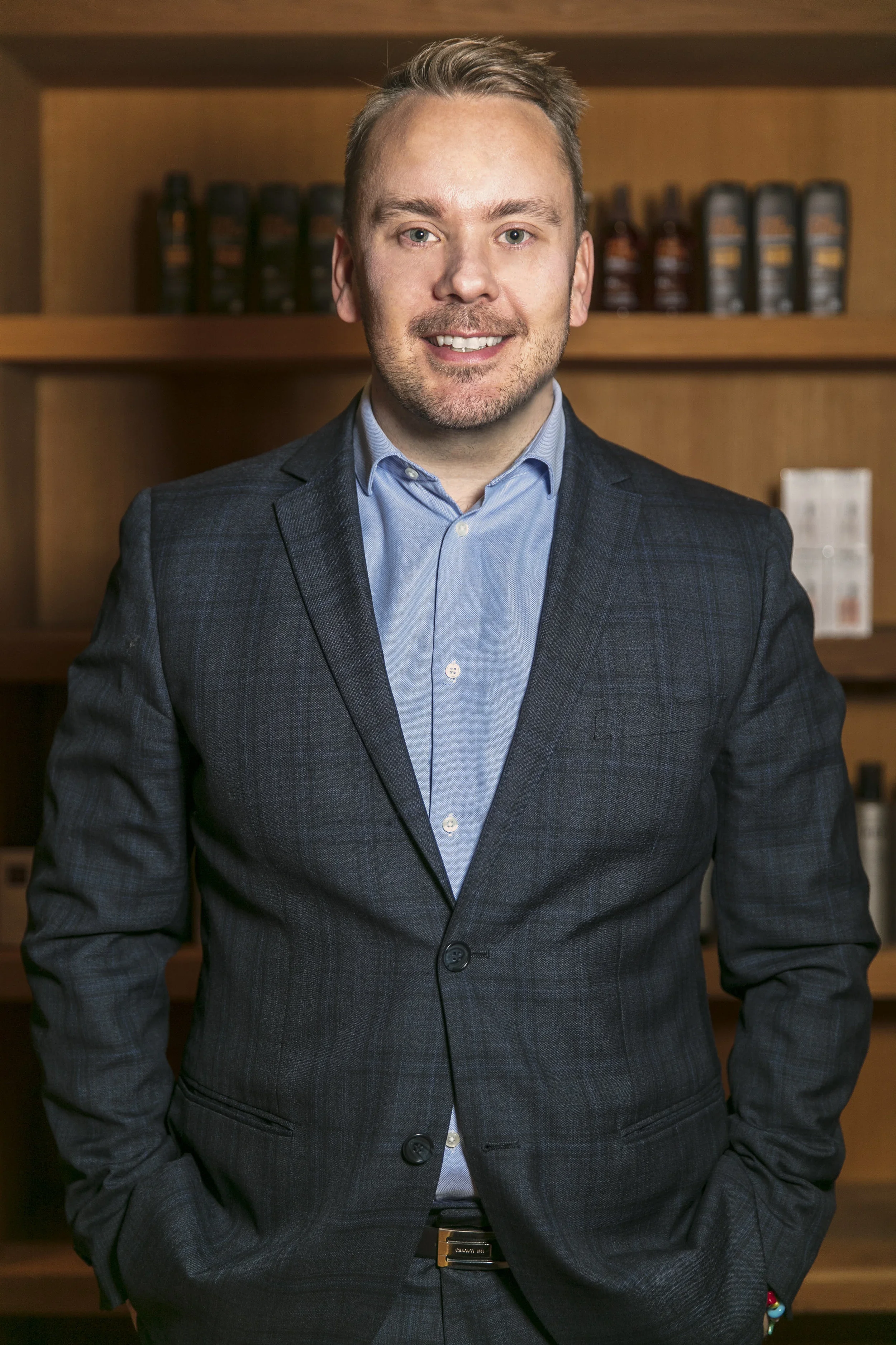 A man in a dark plaid suit and blue dress shirt standing indoors with wooden bookshelves in the background.
