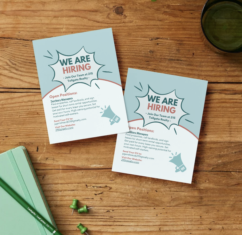 Two light blue flyers on a wooden desk advertise a job opening at 215 Tollegate Realty, with details about open positions and contact information, alongside green push pins, a green notebook, a green pen, and a dark cup.