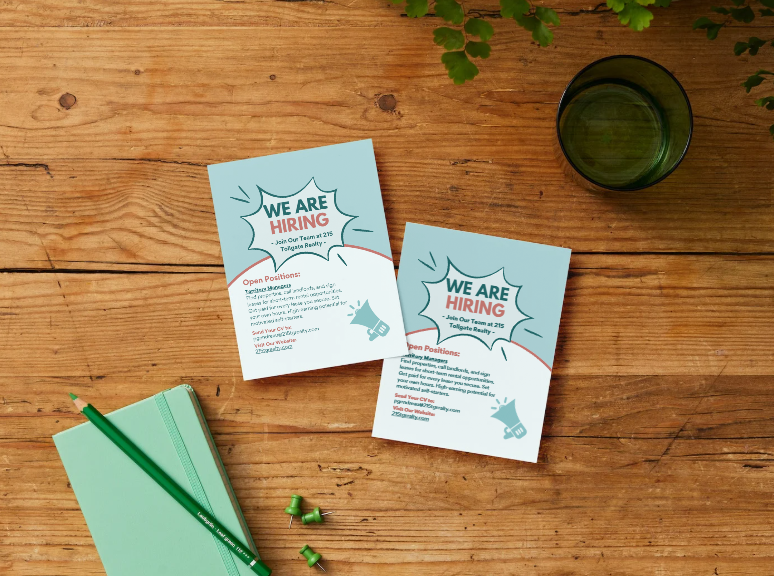 Two recruitment flyers with 'We Are Hiring' headline, on a wooden table next to a mint green notebook with a green pencil, a glass of water, and green push pins.