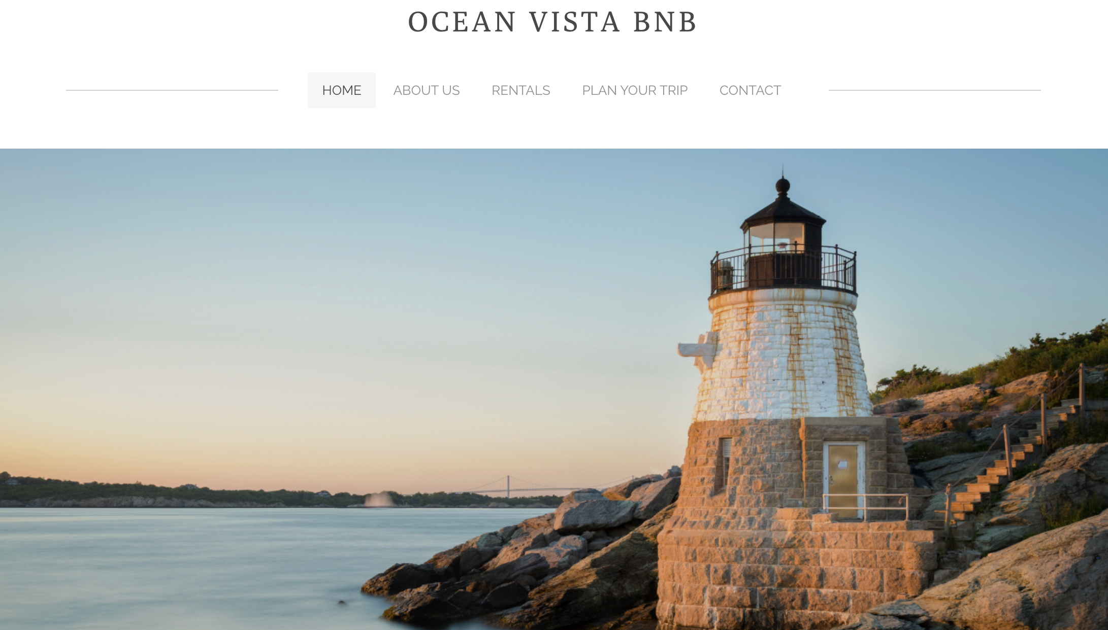 Lighthouse on rocky shoreline during sunset, with water in the foreground and a bridge in the distance.