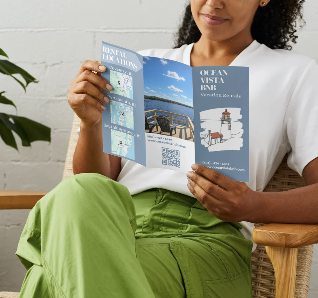 A person sitting on a chair holding a brochure for Ocean Vista BNB vacation rentals, showing maps of rental locations in Rhode Island, a picture of a dock with a lake, and a lighthouse illustration.