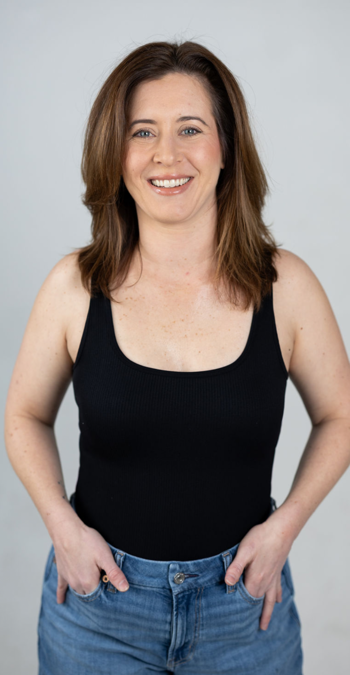 A woman with shoulder-length brown hair smiling, wearing a black tank top and blue jeans, posing against a plain light gray background.