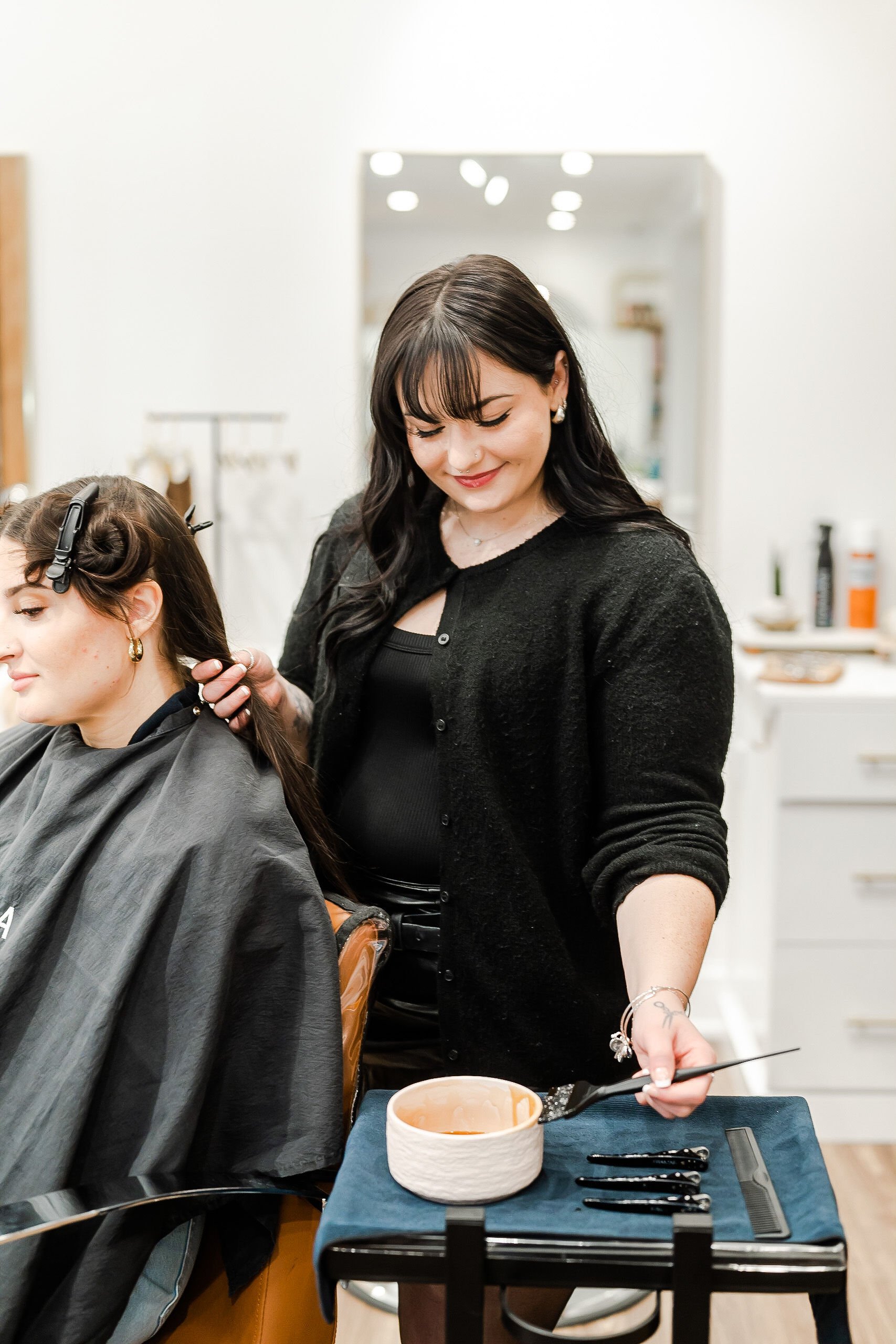 A hairstylist with black hair, in a black cardigan, smiling while applying hair color or treatment to a client in a salon. The client has dark hair, hair clips, and is wearing a black cape. The salon interior is bright with mirrors and shelves in the background.