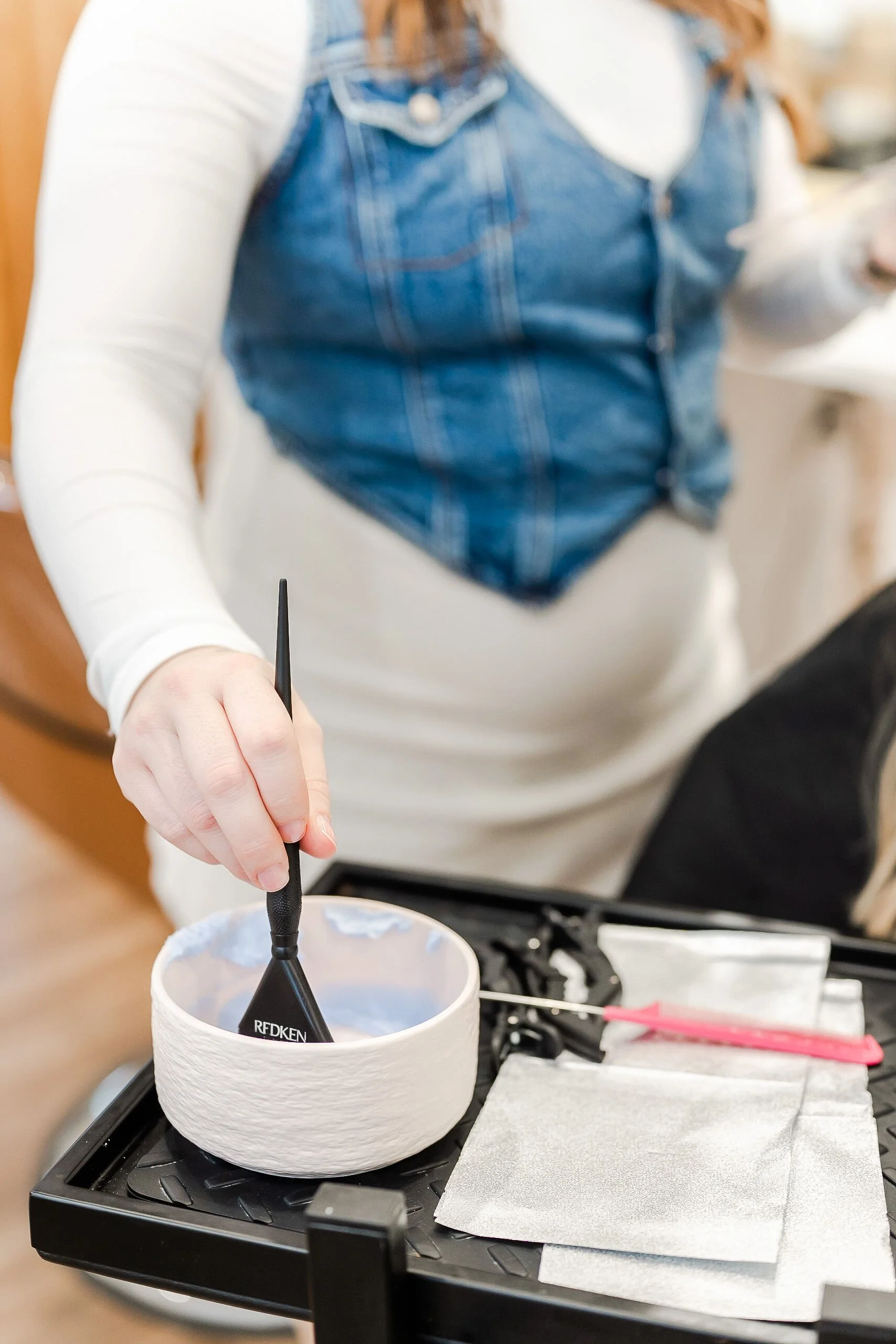 A person with a white long sleeve shirt and denim vest is using a black tool to work on a small white bowl, which is placed on a black tray with salon tools and materials.