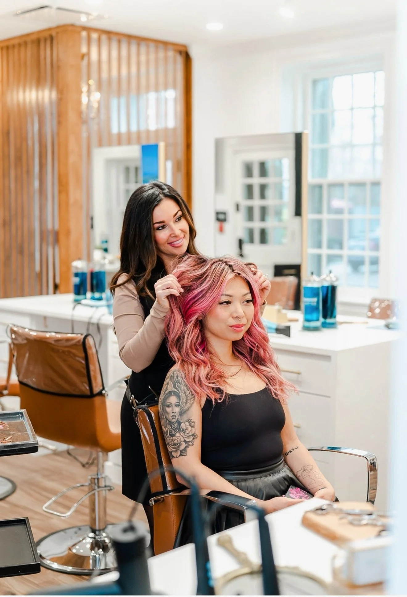 A hairstylist with dark hair styles a woman with pink, wavy hair at a salon. The client wears a black top and has tattoos on her arm, sitting in a salon chair with salon tools and products nearby.