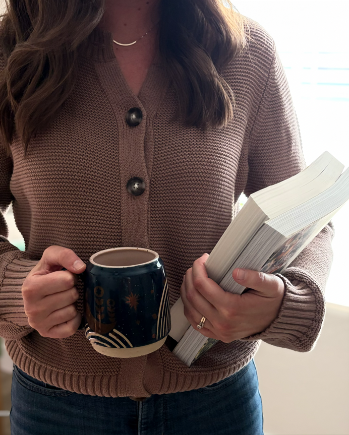 A woman wearing a brown knitted sweater holding a black mug with a starry design in one hand and a thick book in the other.
