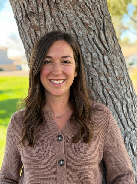 A young woman with long brown hair and a tan sweater, smiling, standing outdoors next to a large tree with a textured bark.