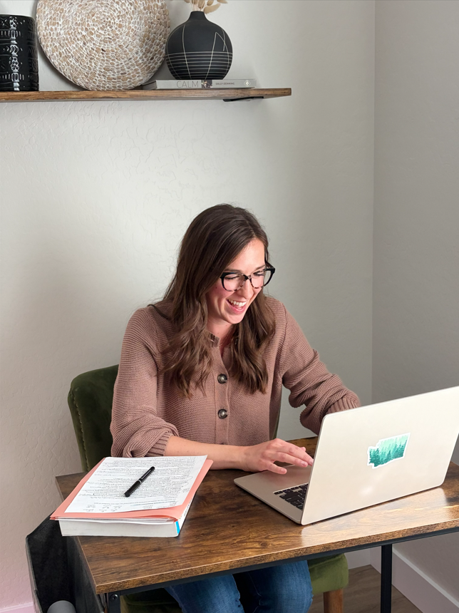 A woman with glasses and long brown hair working on a laptop at a wooden desk, smiling, with a notebook and pen in front of her, in a cozy home office.