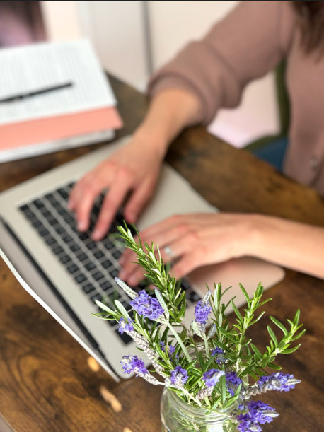 A person working on a laptop with a notebook and vase of purple flowers on a wooden table.