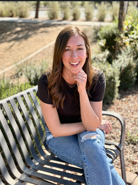 A young woman with long brown hair sitting on a bench outdoors, smiling and wearing a black t-shirt and ripped jeans.