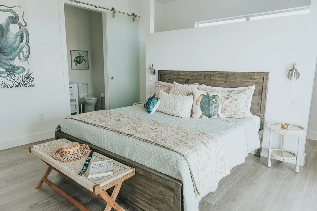 A bedroom with a large bed featuring a wooden headboard, decorated with multiple pillows, a bench at the foot of the bed with a hat and books, and a side table with a small item. There is a wall-mounted reading light and a small bathroom with a toile