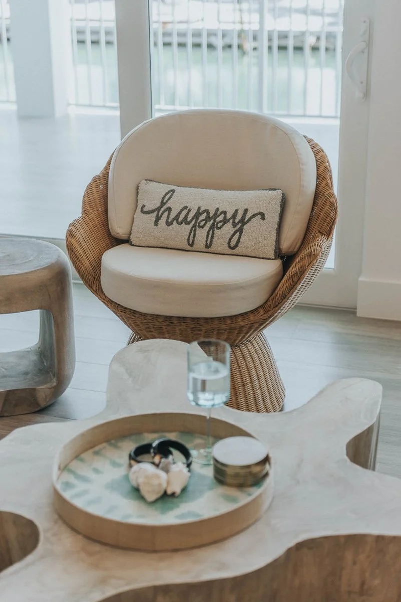 Rattan chair with beige cushion and pillow that says 'happy', glass of water, jewelry, and small decorative items on a wooden table in a bright room with large windows.
