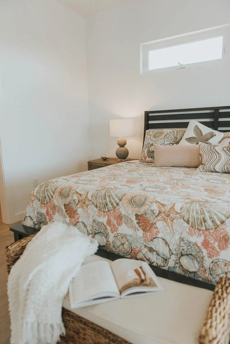 Bedroom with a bed featuring a seashell and starfish patterned quilt, pillows, a nightstand with a lamp, and an open book with a white blanket draped over a wicker bench.
