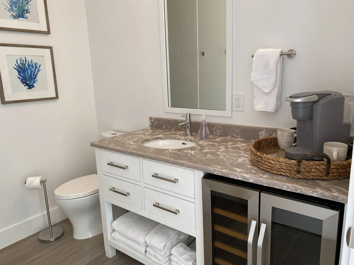 Bathroom with toilet, mirror, vanity, coffee maker, towels, artwork, and glass-doored beverage cooler.