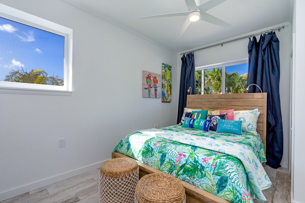 Bedroom with a wooden bed, colorful tropical-themed bedspread, blue curtains, two wicker stools, wall art, and windows showing tree foliage outside.