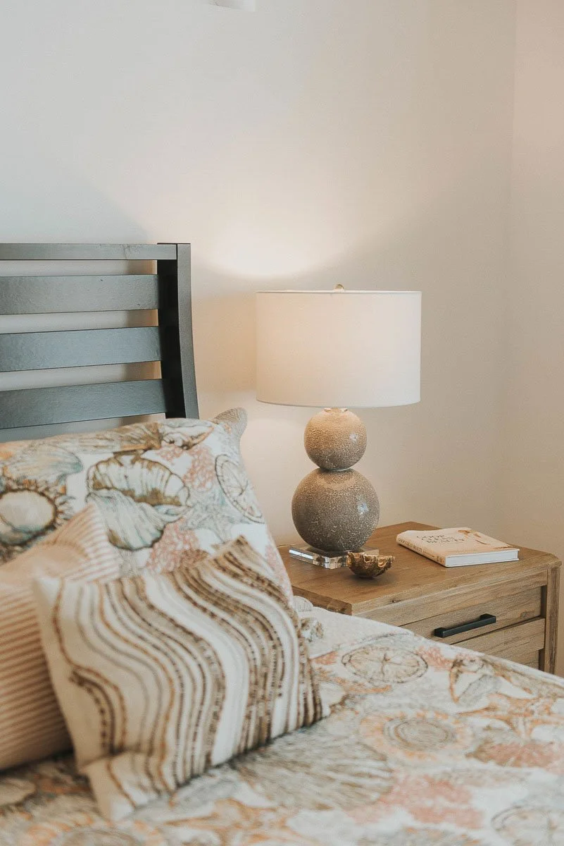 A bedroom scene with a bed that has patterned pillows, a wooden nightstand, a decorative table lamp with a stone base, a closed book, and a small decorative bowl, against plain light-colored walls.