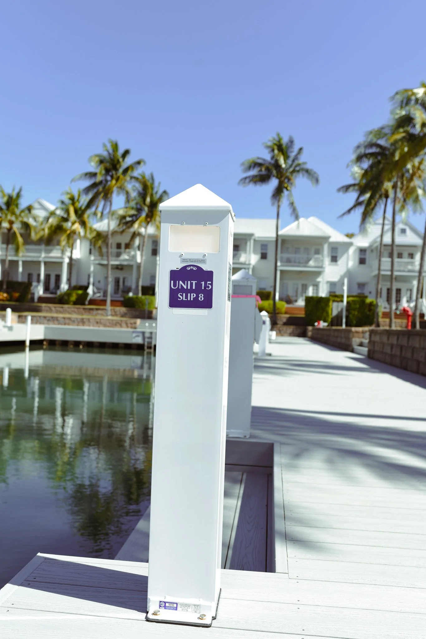 White dock post with purple sign reading "Unit 15 Slip 8" at a marina, with waterfront houses, palm trees, and a clear blue sky in the background.