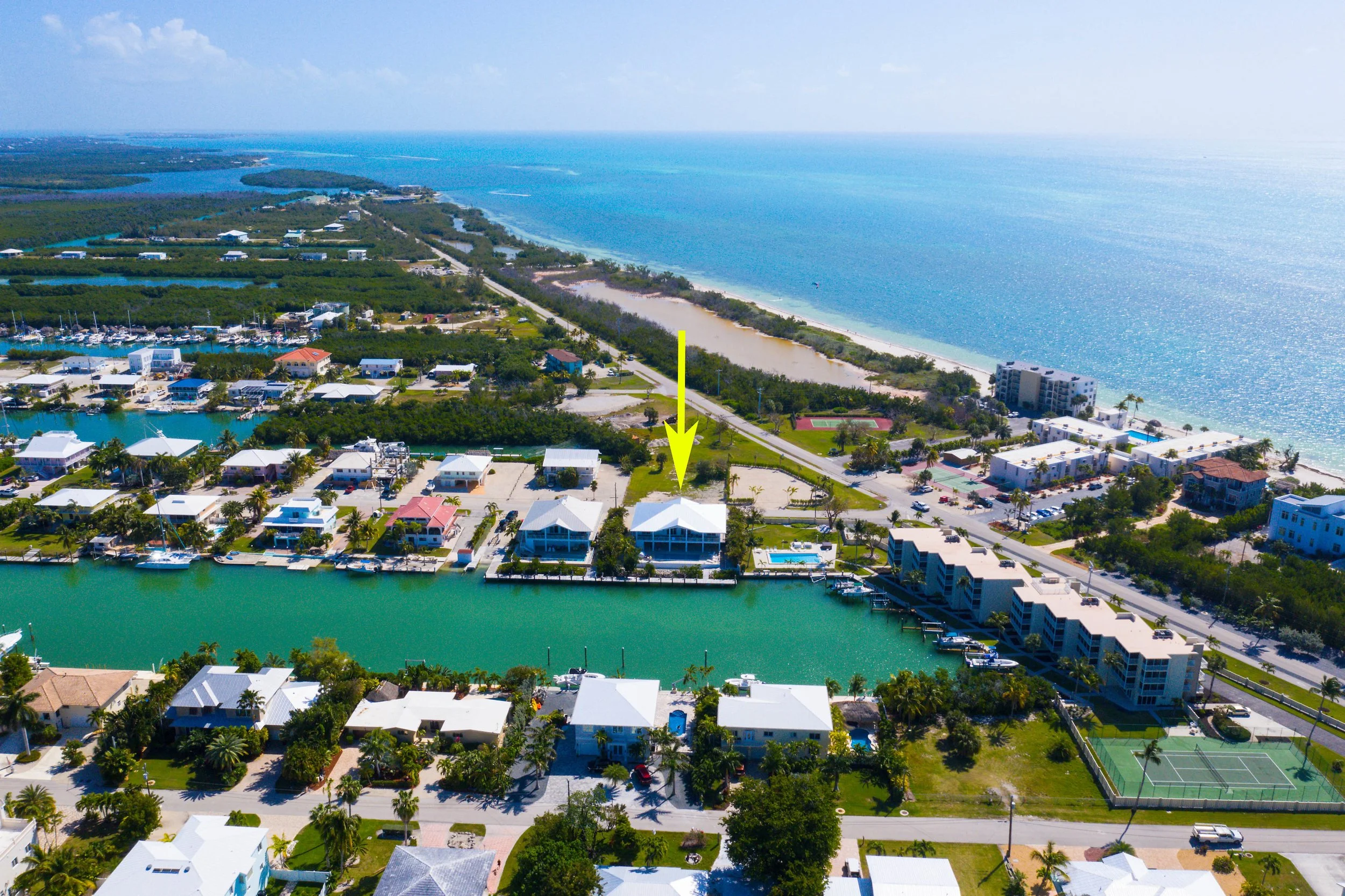 Aerial view of a coastal neighborhood with houses, a canal, and the ocean in the background. A yellow arrow points to a specific house with a white roof near the water, and there are other homes, boats, and tennis courts nearby.