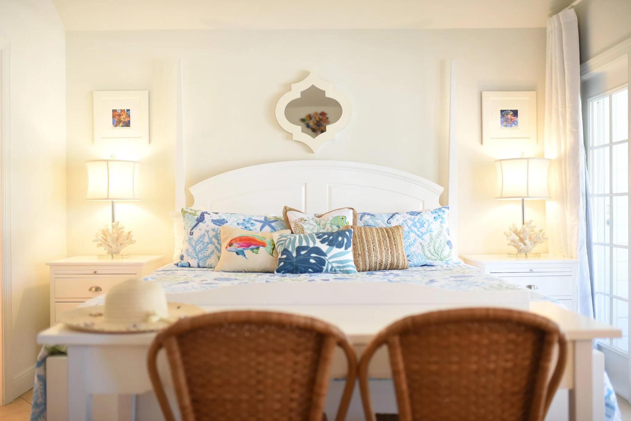 A bedroom with a white bed, decorated with ocean-themed pillows, accompanied by two nightstands with lamps. The room has light-colored walls, an ornate mirror above the bed, and a window with curtains to the right.