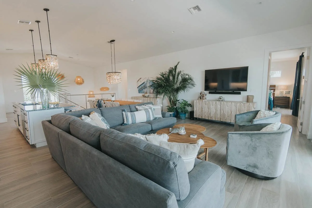 Modern open-concept living room with gray sofas, a large flat-screen TV, decorative plants, wooden coffee tables, and pendant lights. Adjacent to a dining area with a table and chairs, and a bedroom visible through a doorway in the background.