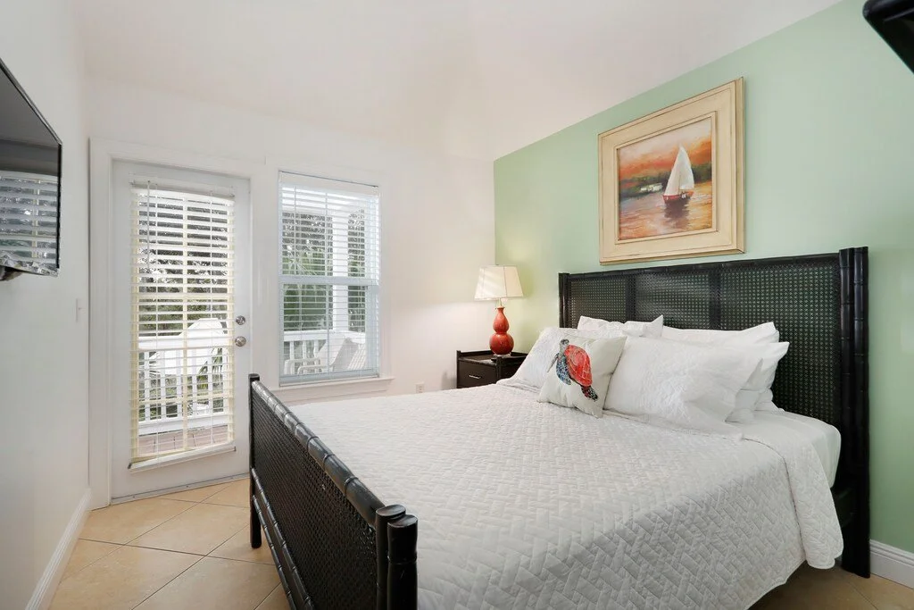 A bedroom with a king-sized bed, white quilt, decorative pillows, a framed sailboat painting above the headboard, a bedside table with a lamp, and a balcony door with blinds leading outside.
