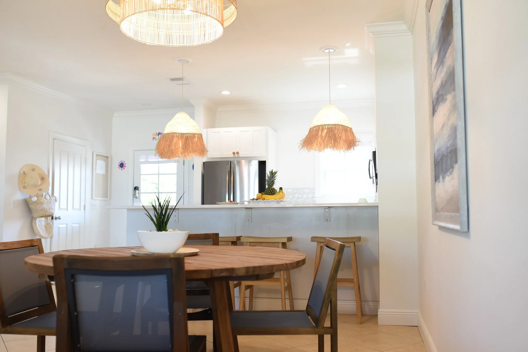 A bright kitchen and dining area with a round wooden table, four chairs, hanging pendant lights with fringed shades, a bowl of fruit, and a pineapple on the counter.