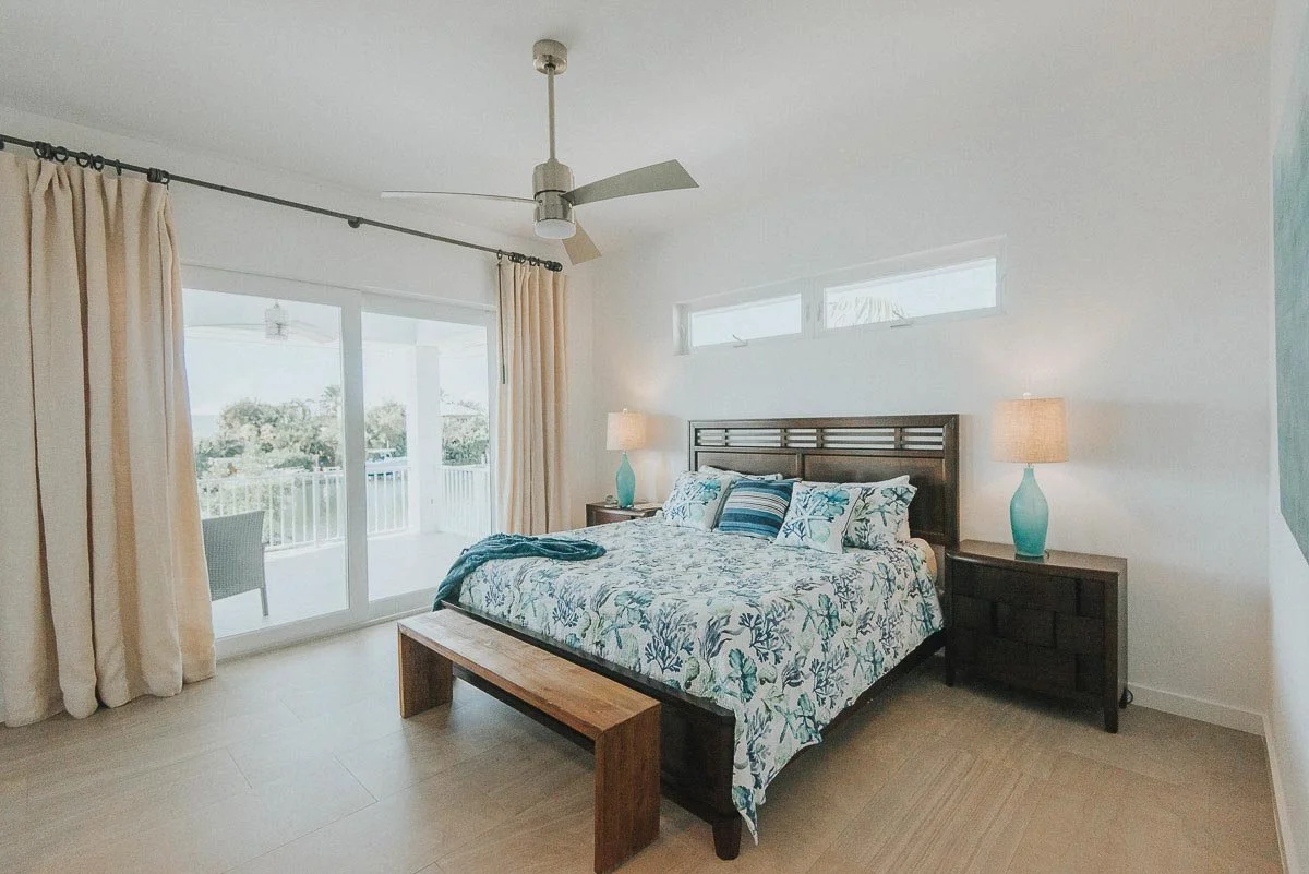 A bedroom with a king-sized bed covered in a blue, white, and green patterned comforter and pillows, two bedside tables with matching blue lamps, a ceiling fan, and sliding glass doors leading to a balcony with outdoor seating, overlooking a green la