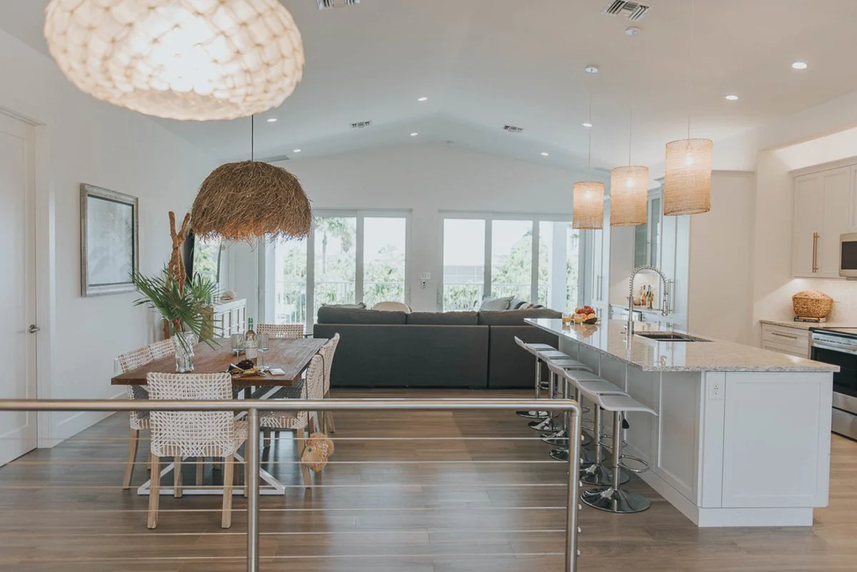Open-concept living room and kitchen in a modern home with white walls, hanging pendant lights, a large kitchen island, bar stools, a dining area, and large windows with a view outside.