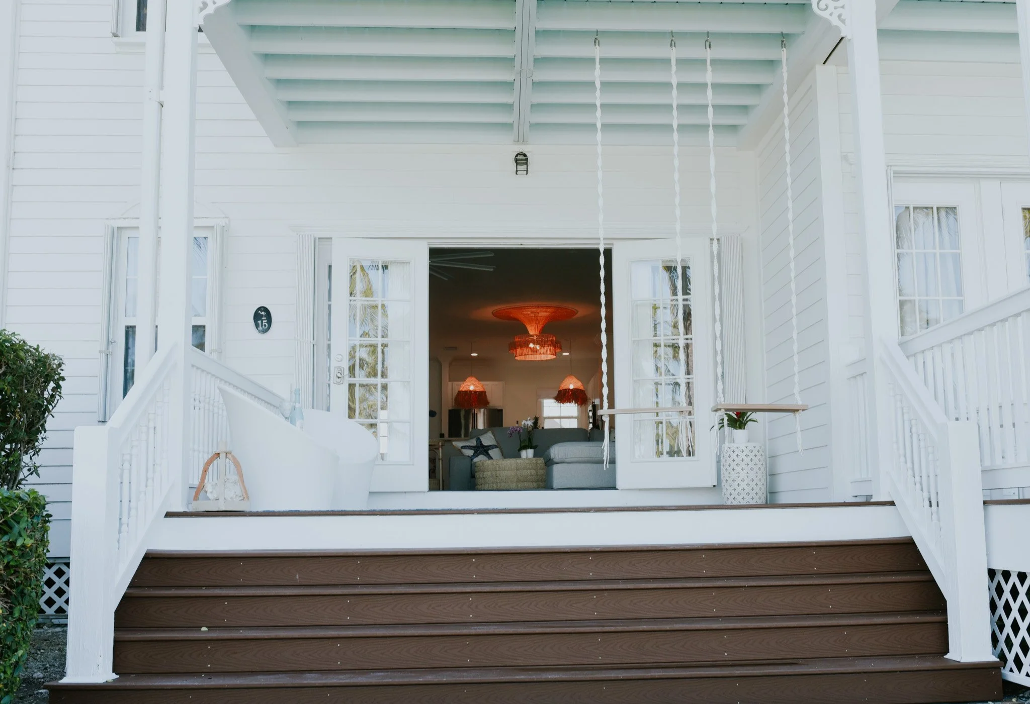 View of a white house with open glass doors leading to a patio with brown stairs. Inside, hanging orange chandeliers illuminate a living room with a sofa and lamps.