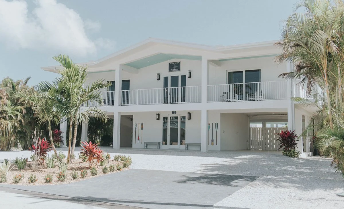 White two-story house with balcony, surrounded by tropical plants and palm trees, under a partly cloudy sky.