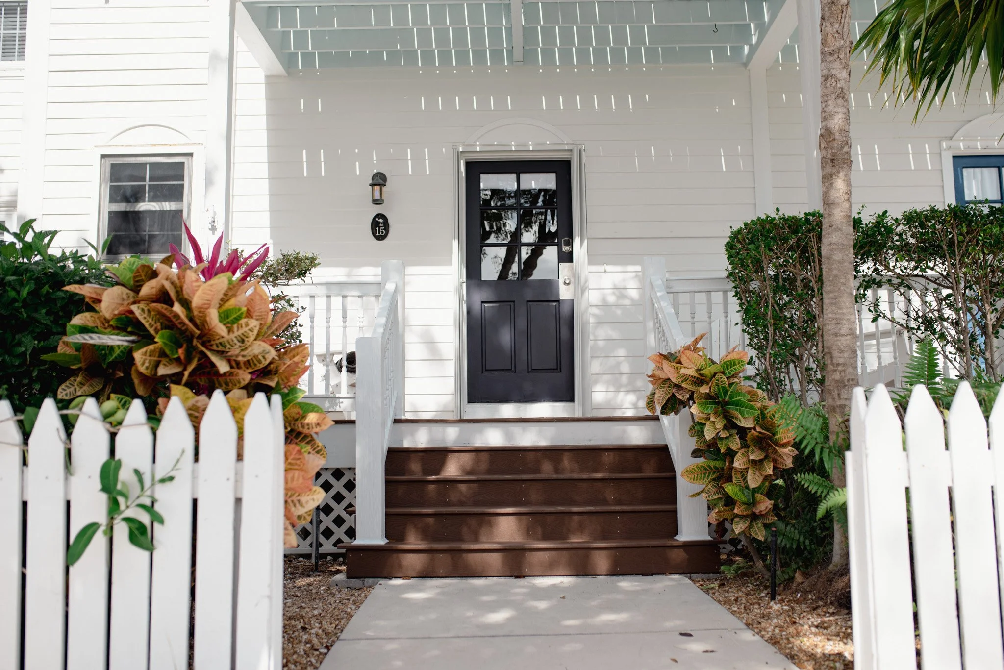 Front view of a white house with black door, small porch with wooden steps, surrounded by plants and a white picket fence.